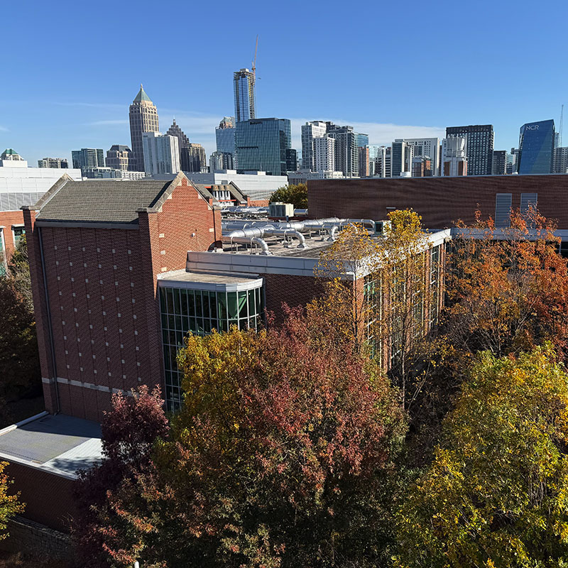 Aerial view of a brick academic building surrounded by autumn trees, with the Atlanta skyline and modern high-rise buildings in the background under a clear blue sky.