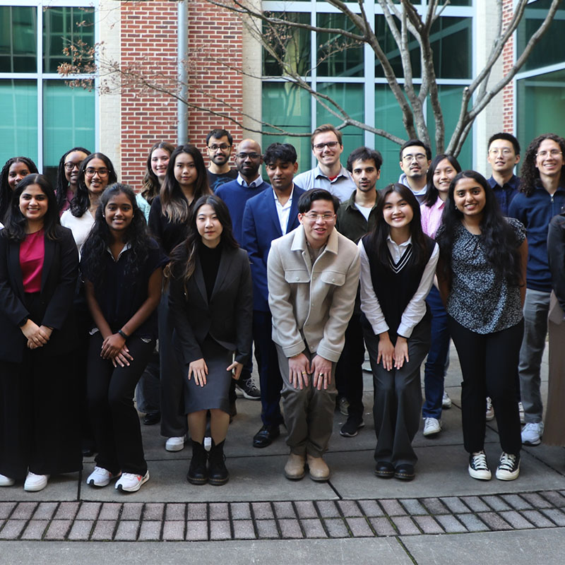 A large group of students standing together outdoors in a courtyard, posing for a group photo in front of a building with brick walls and large windows.