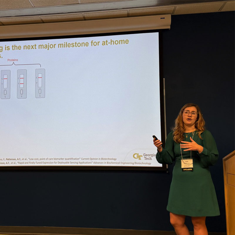 A presenter in a green dress speaks beside a large projection screen displaying a slide about at‑home diagnostic testing at a Georgia Tech event.