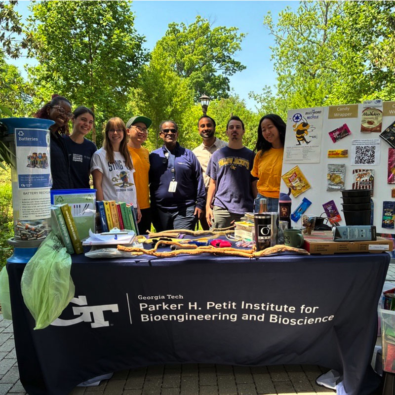 Group of people standing behind an outdoor information table covered with a navy blue tablecloth labeled “Georgia Tech Parker H. Petit Institute for Bioengineering and Bioscience.” The table displays books, containers, and examples of recyclable items, including batteries and candy wrappers, with posters explaining recycling. Trees and a paved walkway are visible in the background on a sunny day.