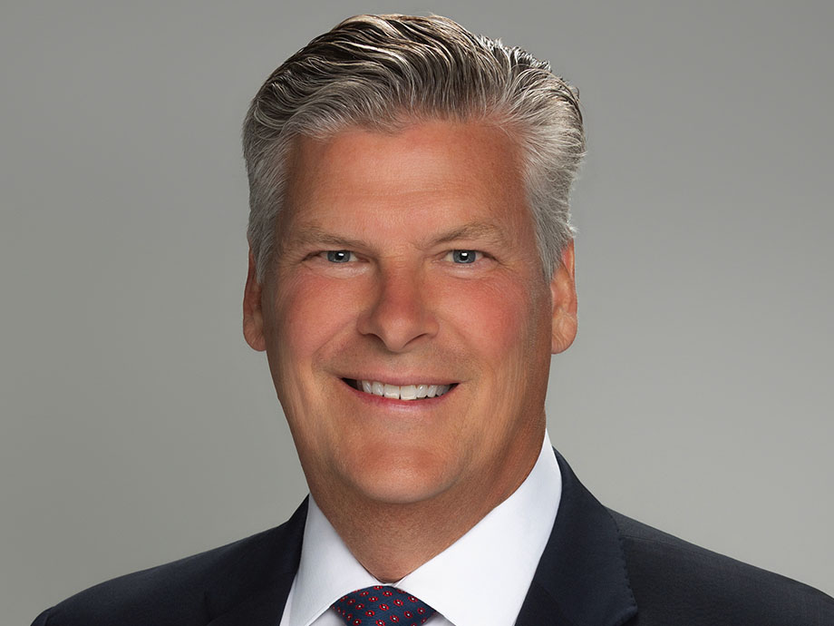 Studio headshot of a white man with short, neatly styled gray hair, wearing a dark suit jacket, white dress shirt, and patterned tie. The person is photographed from the chest up against a smooth light gray background, facing the camera with even, professional lighting.