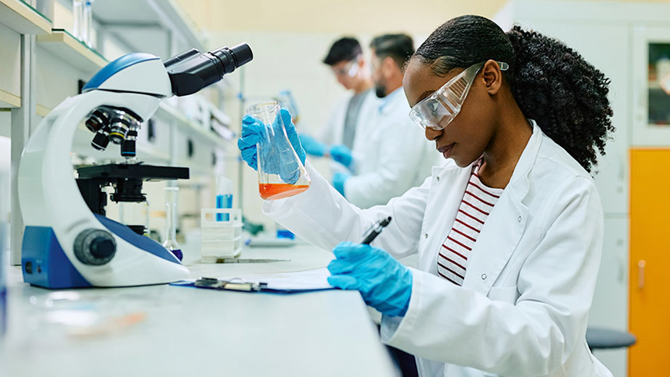 A researcher wearing gloves examines an orange liquid sample in a laboratory while working beside a microscope and lab bench equipment.