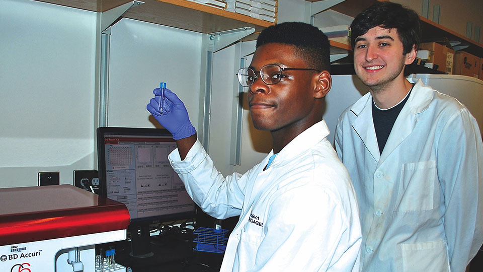 Two people in lab coats working in a laboratory, with one holding up a small sample vial near a computer displaying scientific data.