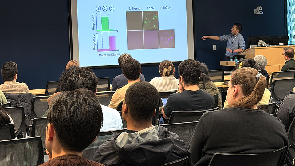 A group of people listen as a man gives a talk at the front of a large seminar room while pointing at a presentation on a projector screen.