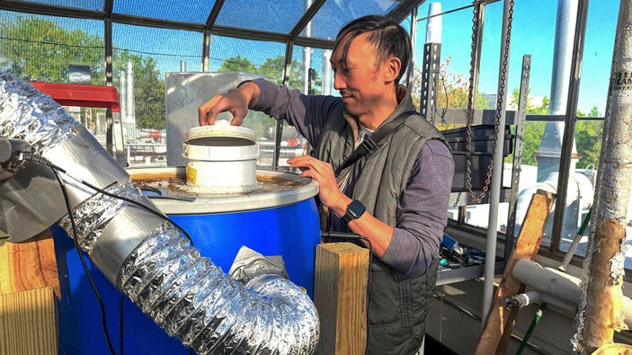 A male researcher opens the top of a blue barrel that is part of a composting system inside a greenhouse