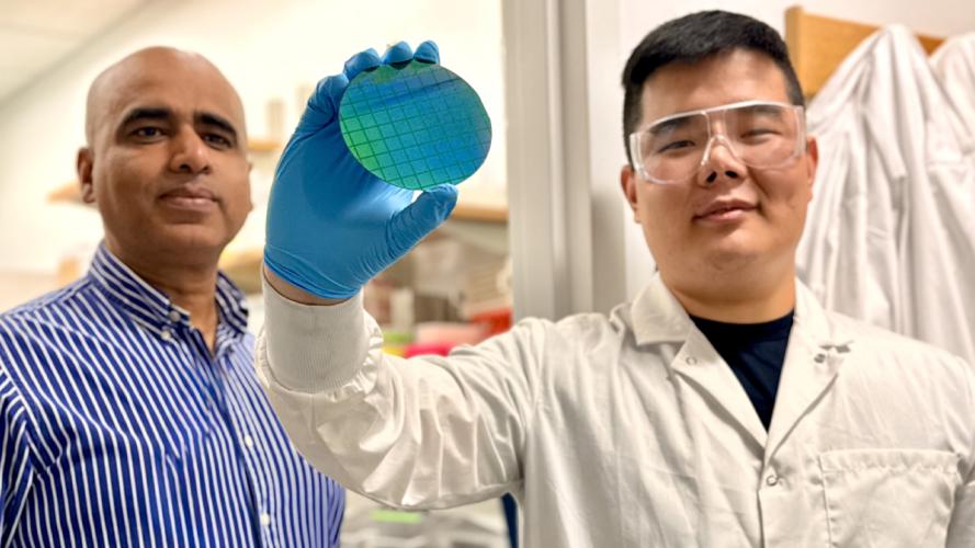Two researchers in a laboratory examine a blue-green silicon wafer. One, wearing a lab coat, safety goggles, and a blue glove, holds the wafer up for inspection while the other stands beside them. Laboratory equipment is visible in the background.