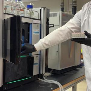 A man wearing latex gloves and a white lab coat puts a tray of samples into a mass spectrometry machine.